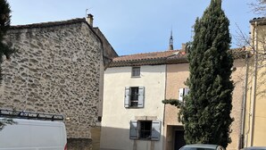 A narrow street with parked cars, a white van, and a building with a stone facade and a tiled roof.