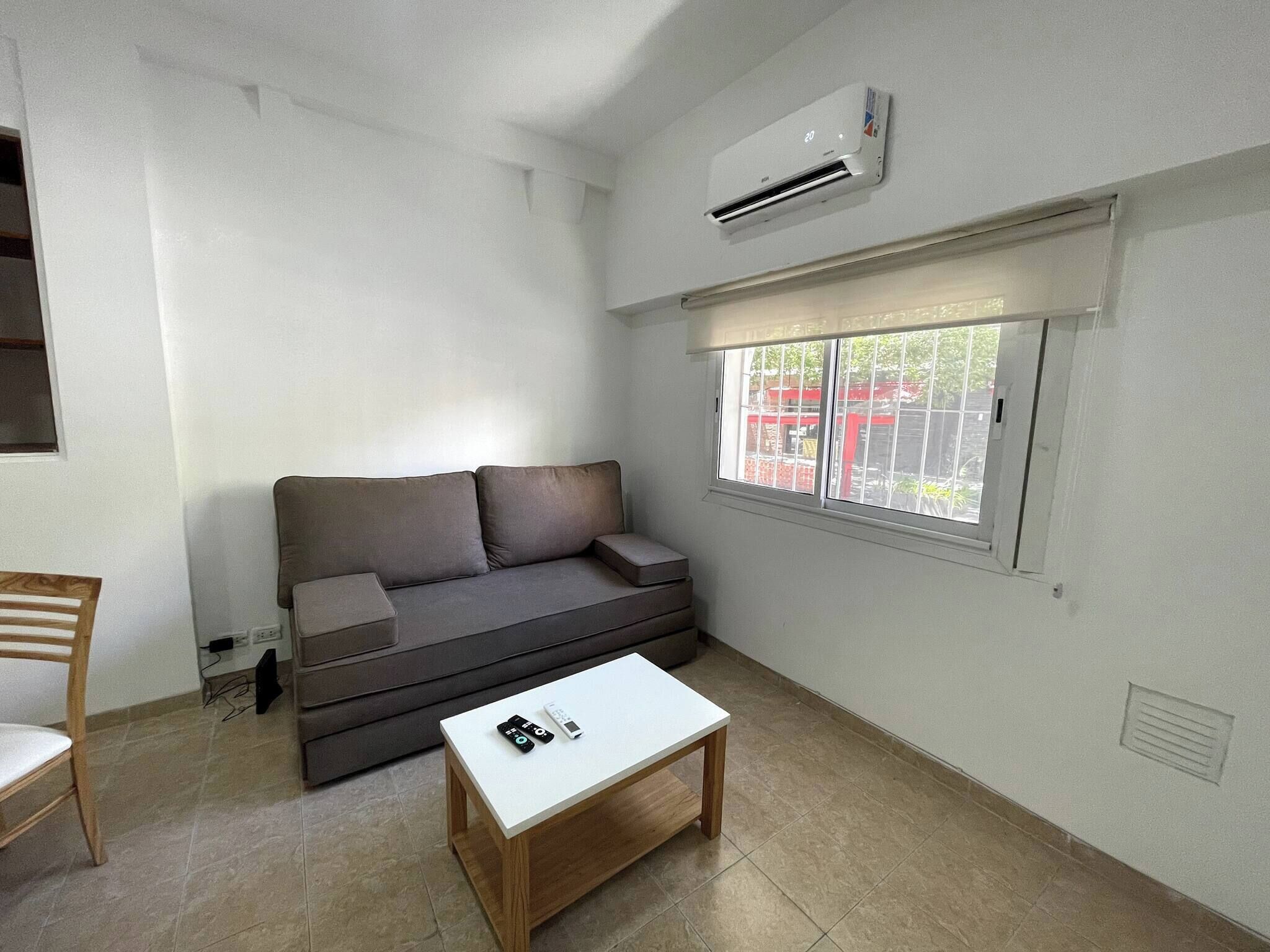 Una sala de estar moderna con un sofá gris, una mesa de centro blanca y un ventanal con vistas a un edificio rojo.