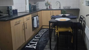 A compact kitchen with wooden cabinets, a black countertop, a dining table with chairs, a black and white rug, and a wall decoration. - Glams 2beds luxury stylish apartment (West Midlands)