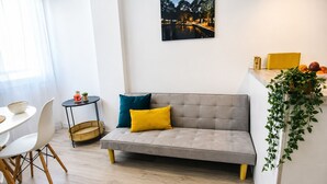 Ein modernes Wohnzimmer mit Sofa, Esstisch, Stühlen, einem kleinen Beistelltisch, einem Blumentopf und einem gerahmten Bild an der Wand.
