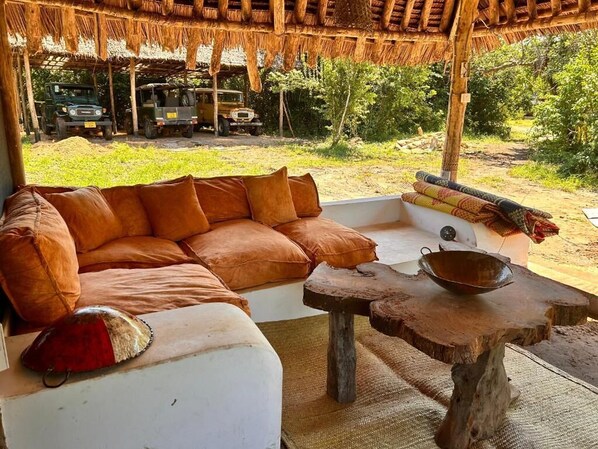 A thatched-roof outdoor seating area with a sofa and a table, surrounded by greenery and vehicles in the background. - Kwa Simba Lodge (Pangani)
