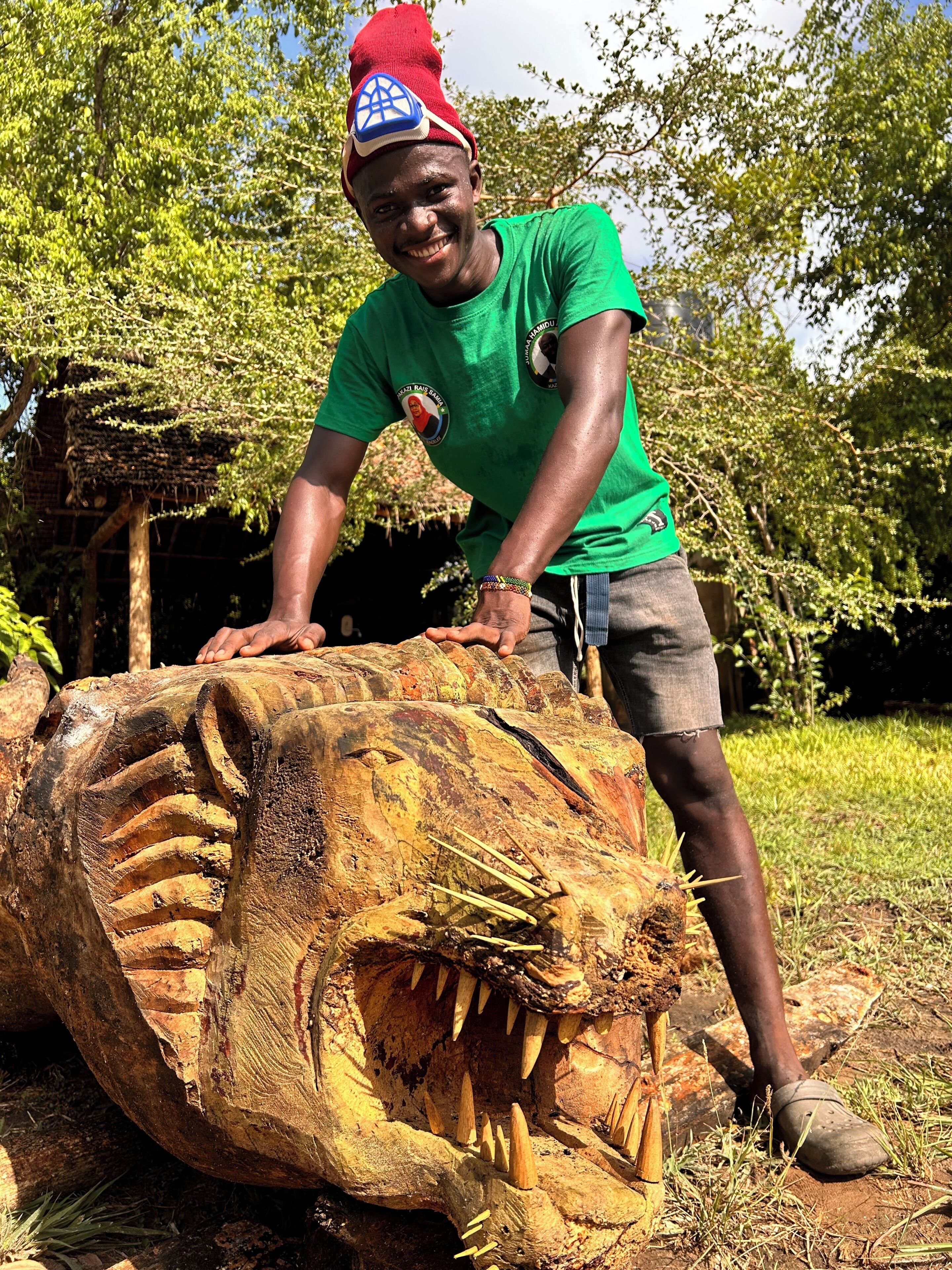 A person in a green shirt and red hat standing next to a large wooden sculpture of a dinosaur.