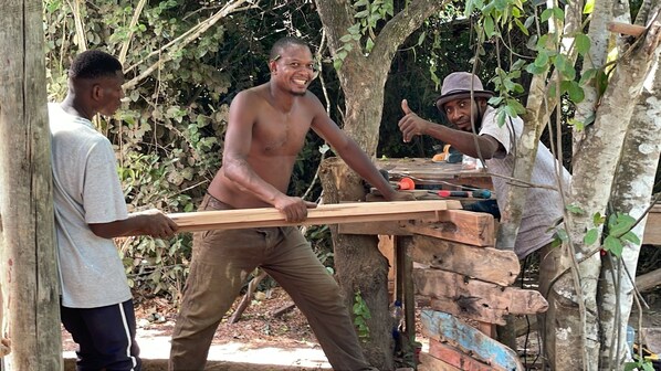 Tres personas realizando trabajos manuales, posiblemente carpintería, al aire libre, con árboles y una estructura de madera improvisada al fondo.