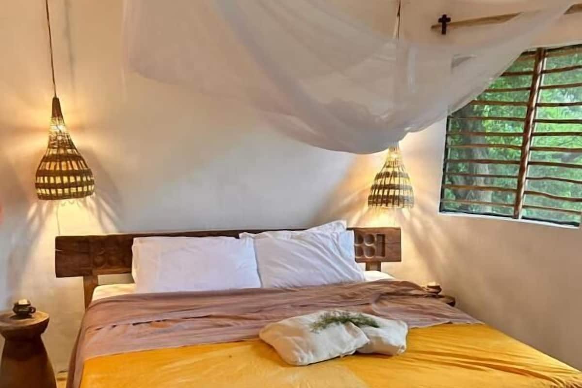 Una cama con colcha amarilla, almohadas blancas y dosel. Hay dos lámparas fijadas a la pared a ambos lados de la cama.