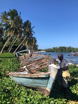 Un homme se tient à côté d’un bateau partiellement démonté, sur une pelouse, avec des palmiers en arrière-plan.