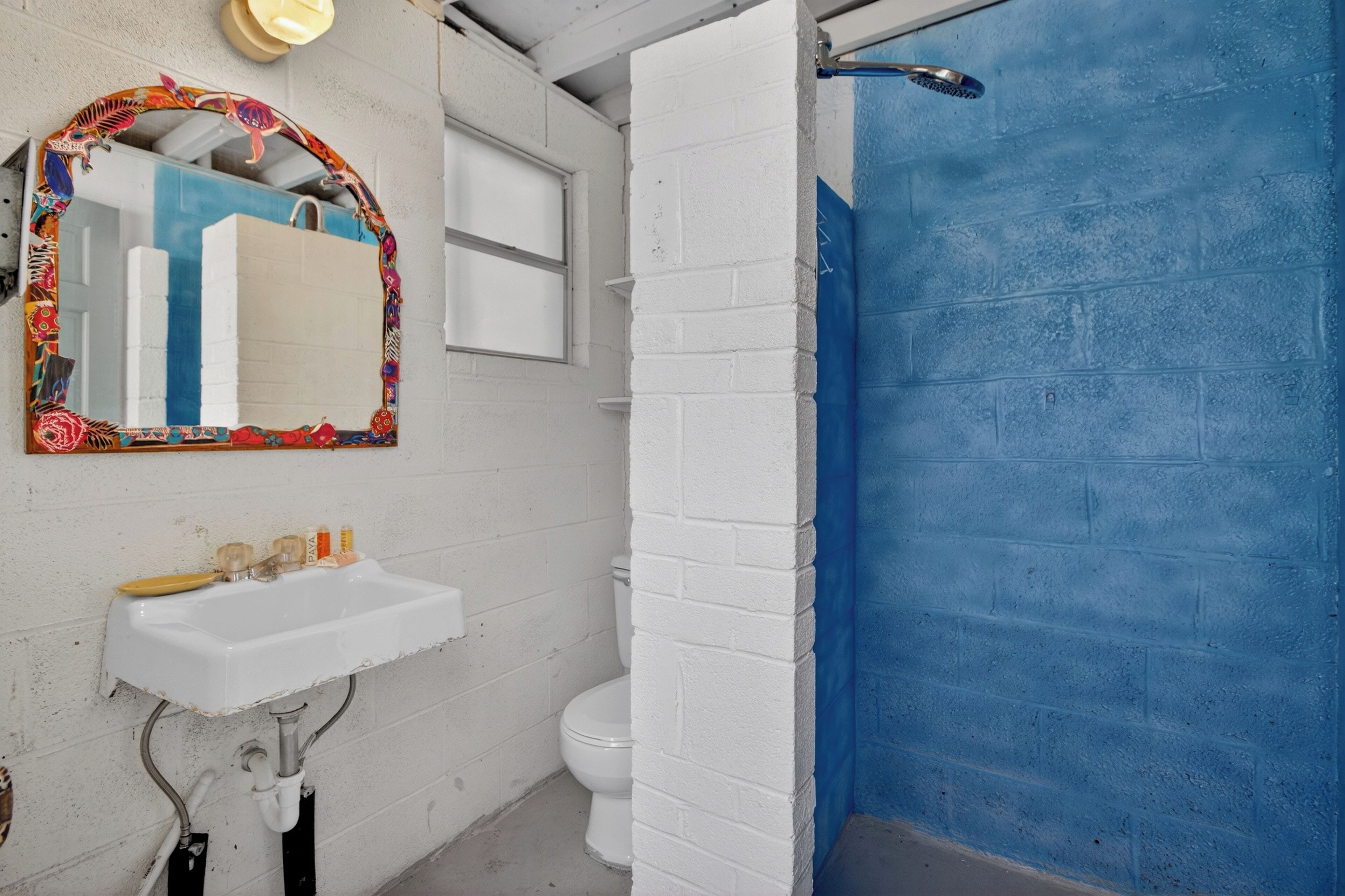 A bathroom with a white sink, a mirror with a colorful frame, a toilet, and a shower area with a blue wall.