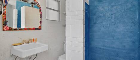 A bathroom with a white sink, a mirror with a colorful frame, a toilet, and a shower area with a blue wall.