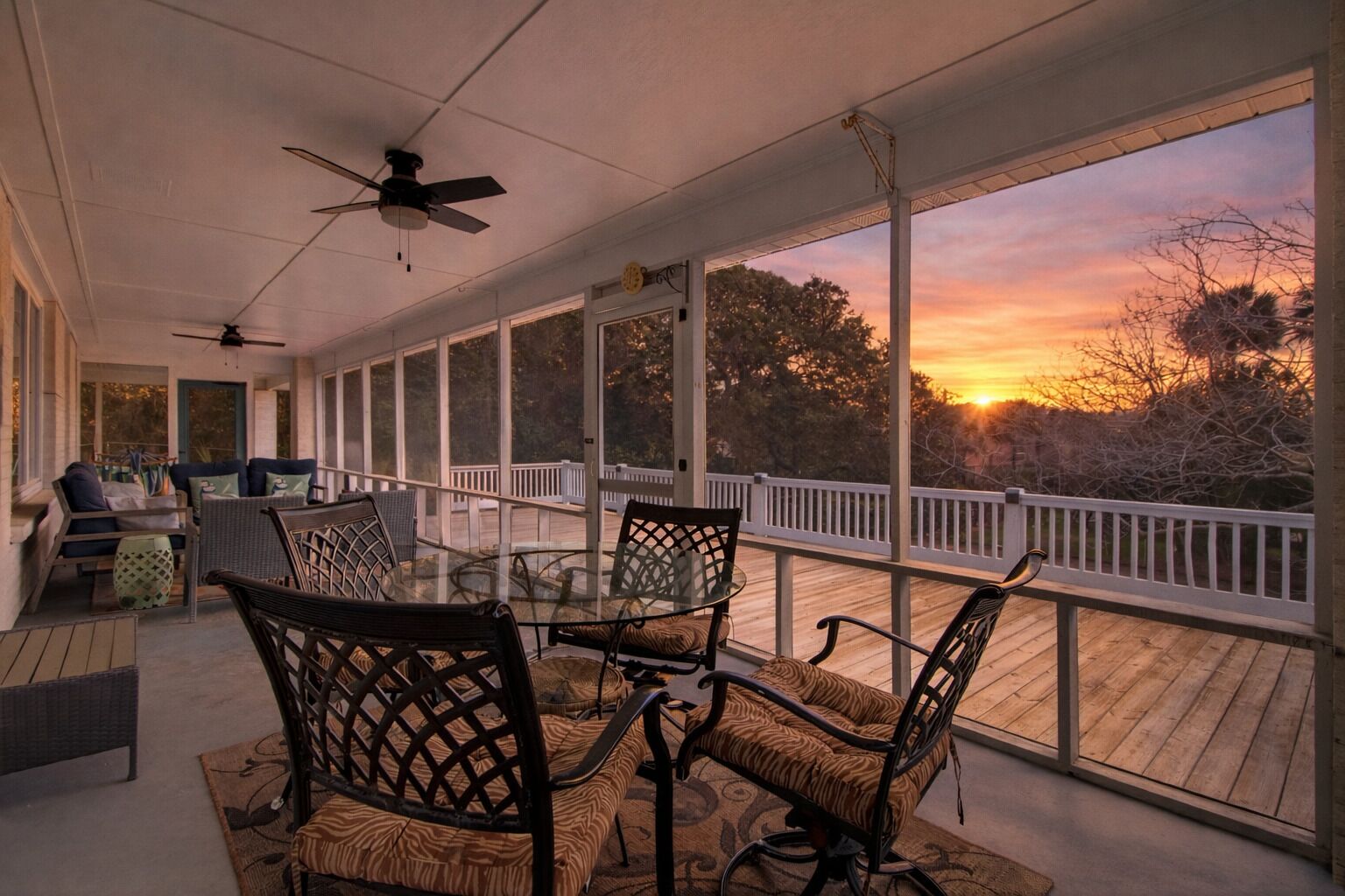 Une terrasse couverte avec une table et des chaises en verre, un ventilateur de plafond et une vue sur le coucher du soleil.