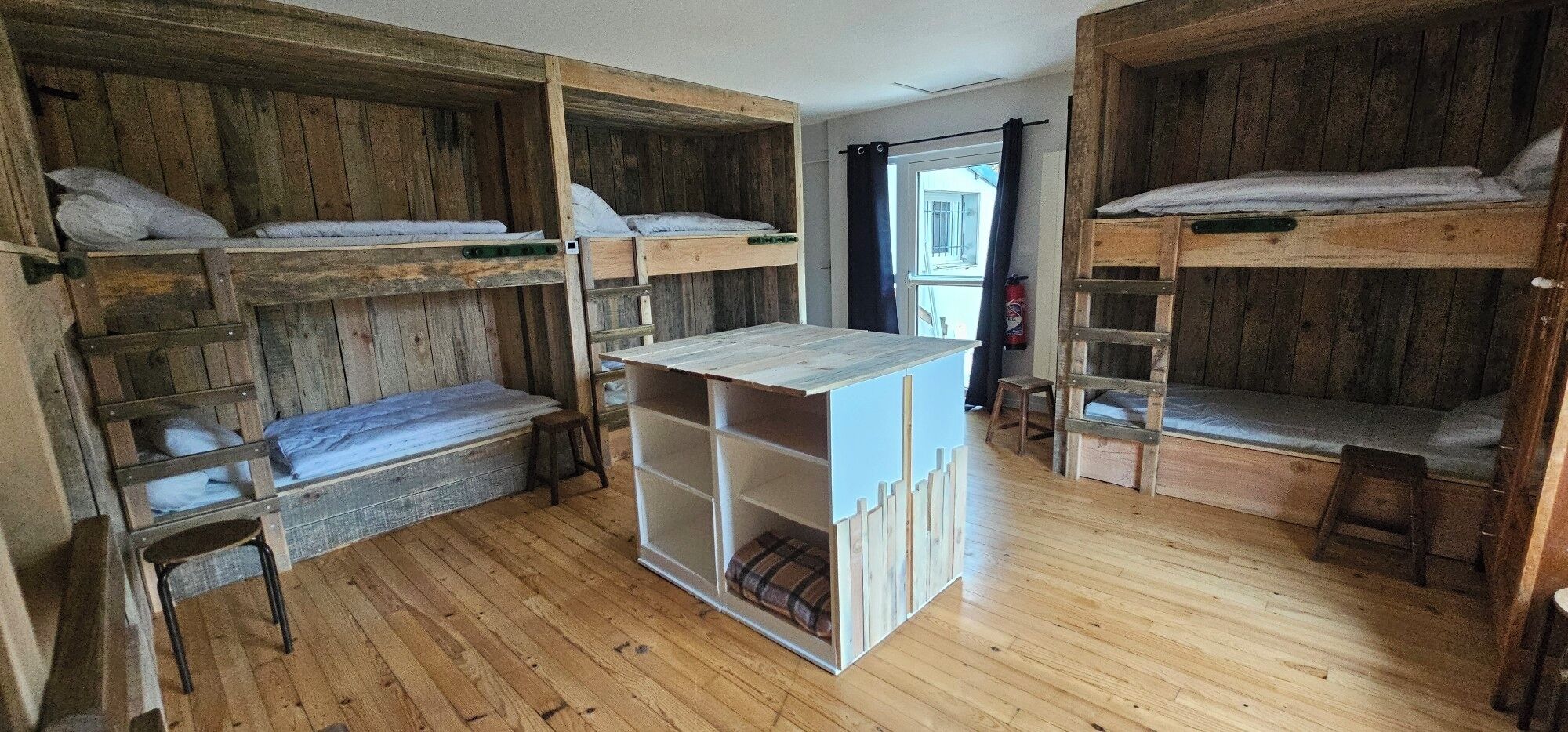 A dormitory room with bunk beds, a central wooden table, and a window with a view.