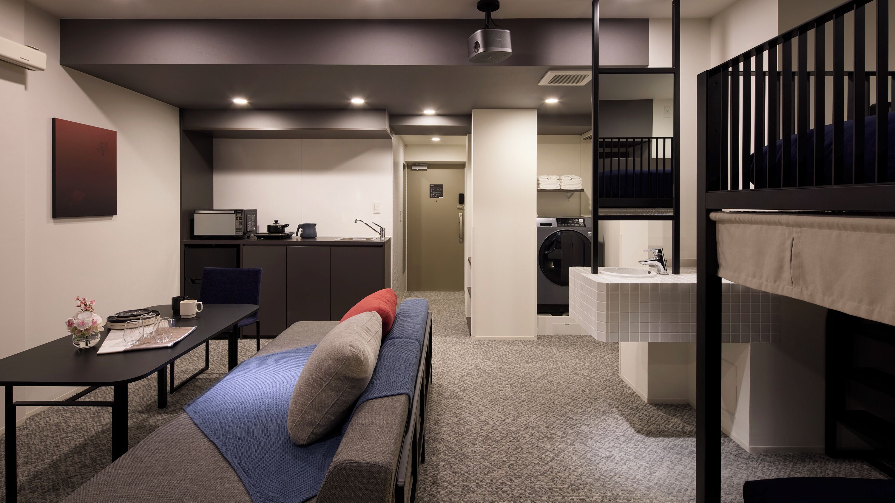 A compact hotel room with a kitchenette, a dining area, a sofa, a small table, and a bathroom with a sink and toilet.