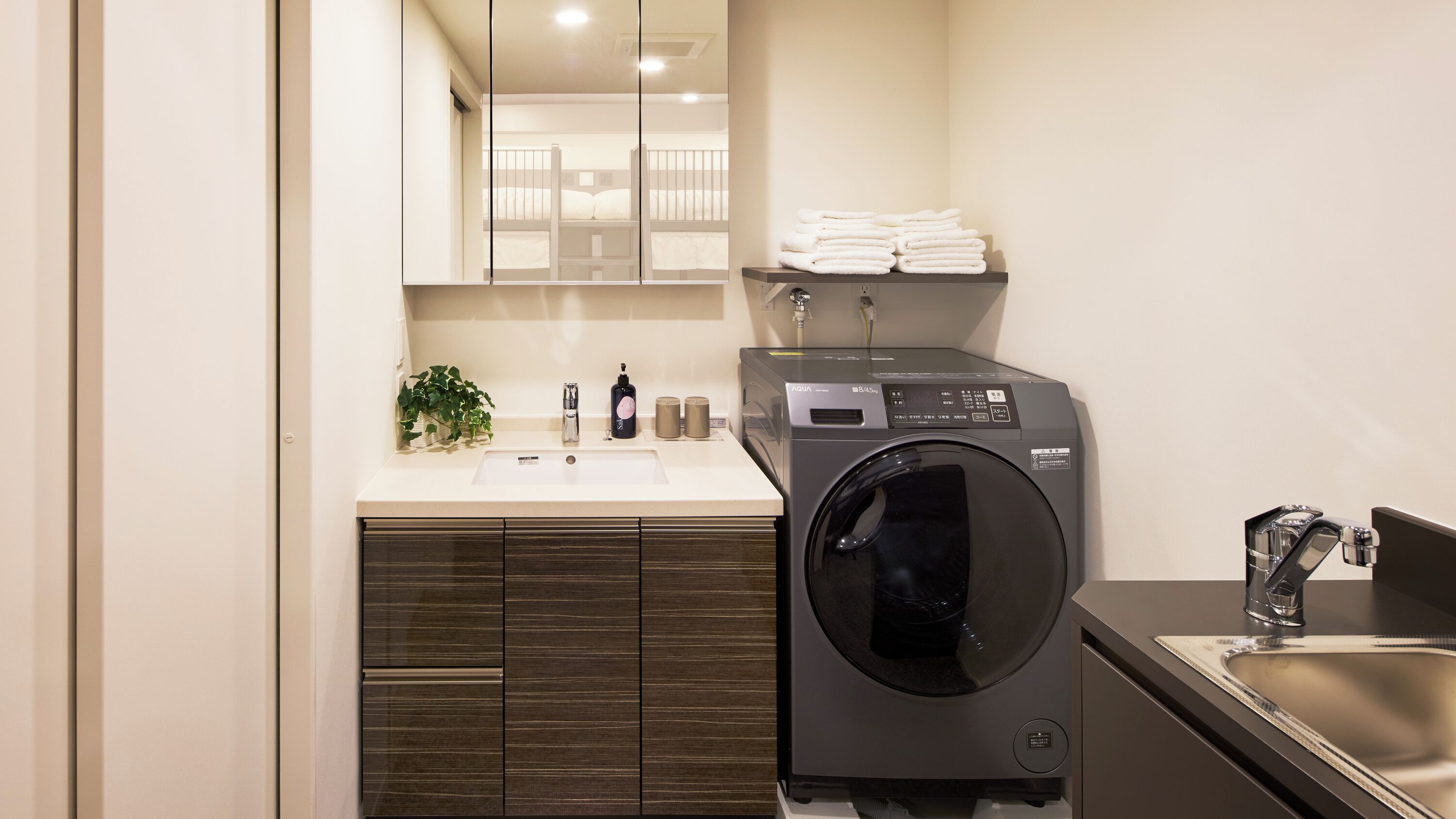 A compact hotel bathroom with a washing machine, a sink, and a mirror.