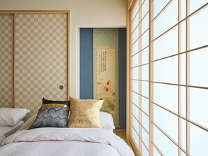 Perinteinen japanilaistyylinen makuuhuone, jossa on tatamimatto, shoji-liukuovi ja useilla tyynyillä varustettu sänky.