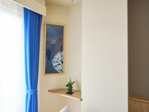 Huoneessa on ikkuna, joka on peitetty sinisillä verhoilla, seinällä on kehystetty taideteos ja seinällä on myös pieni puinen hylly, jonka päällä on ruukussa oleva kasvi.