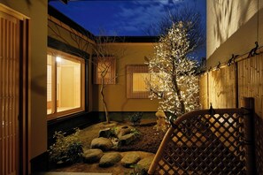 Perinteinen japanilainen puutarha auringonlaskun aikaan, jossa on puinen aita, pieni valoilla koristeltu puu ja kivestä tehty polku.
