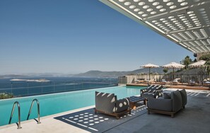 Ein Poolbereich mit Liegestühlen und Sonnenschirmen, mit Blick auf das klare, blaue Meer und entfernte Berge.