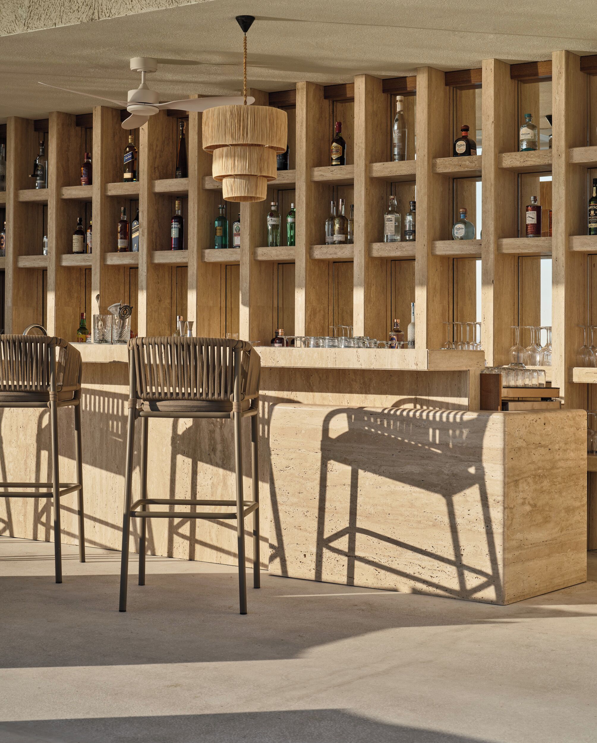 A bar area with wooden shelves stocked with bottles, a hanging light fixture, and metal bar stools.