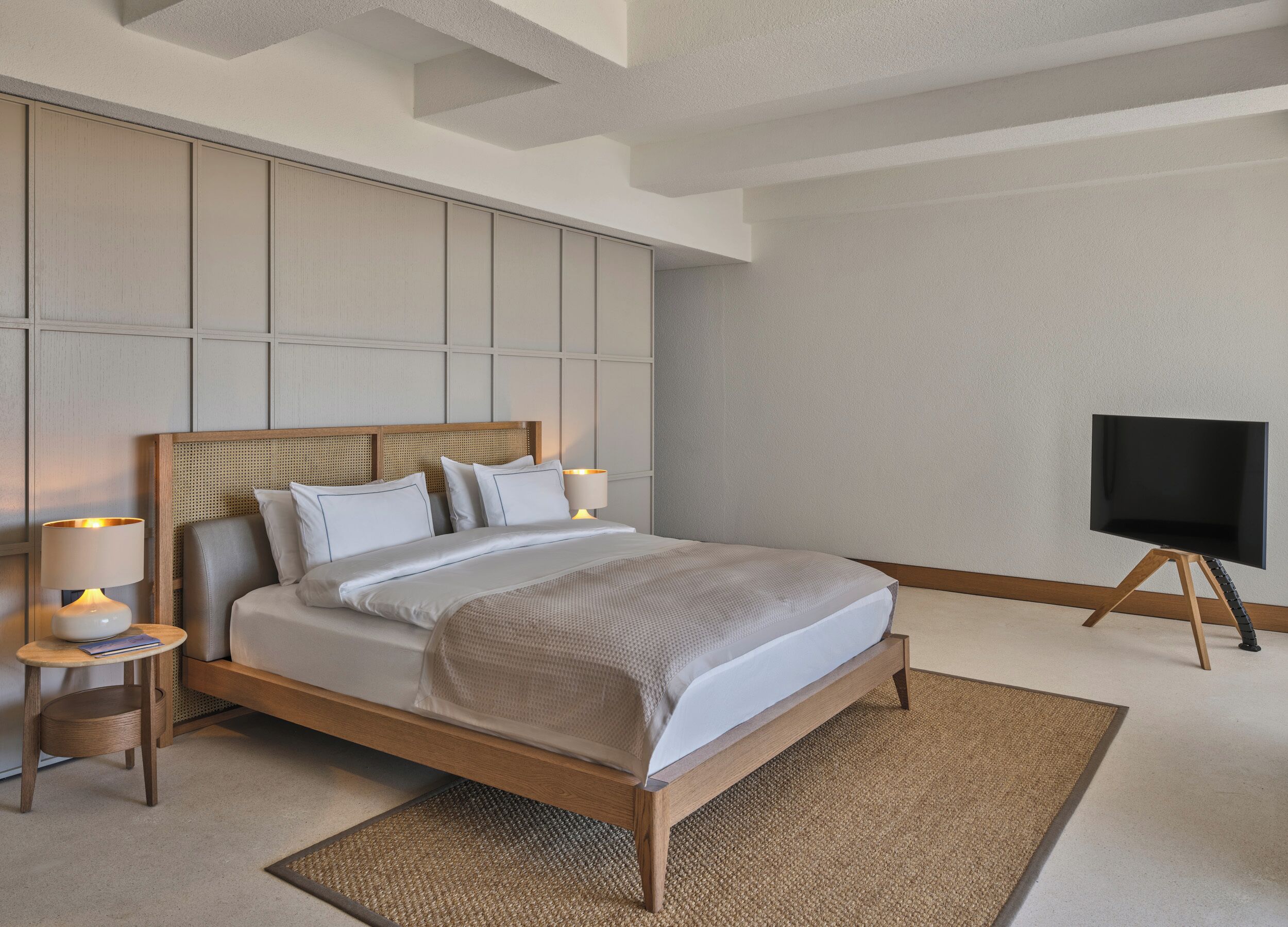 A neatly arranged bedroom with a bed, bedside lamps, a flat-screen TV, and a woven rug.