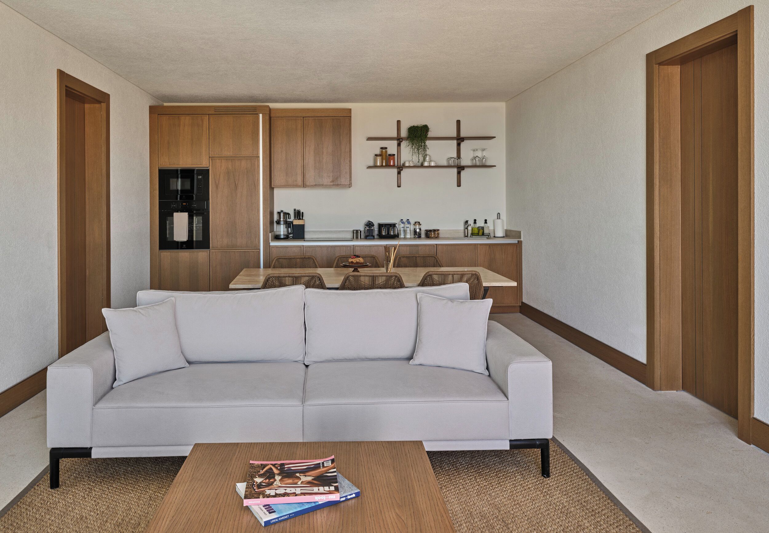 A modern living room with a white sofa, a coffee table with magazines, a kitchen in the background with wooden cabinets and a plant on the wall.