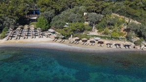 An aerial view of a beach with sun loungers and umbrellas, surrounded by trees and a building with a terrace. - WAM by Karma (Bodrum)