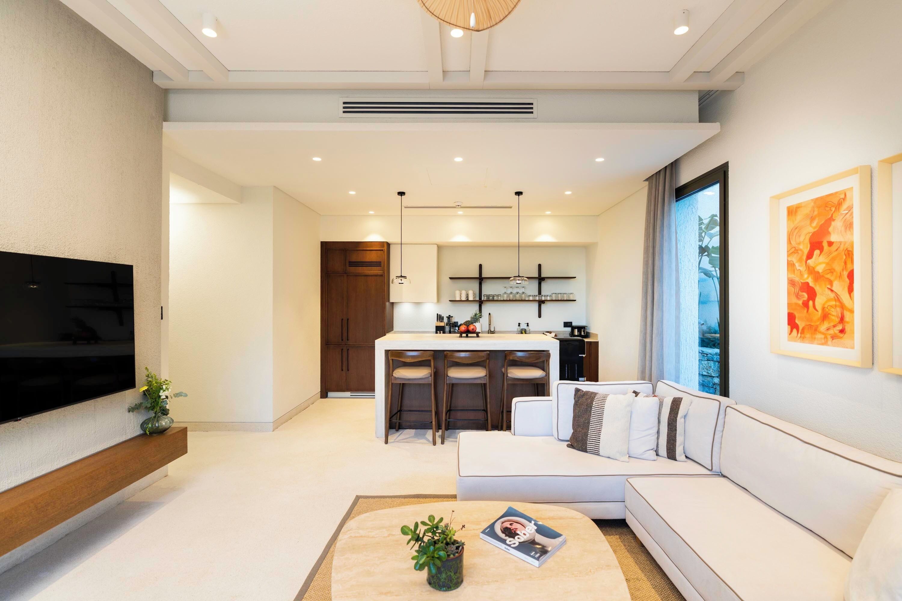 A modern living room with a sofa, a coffee table, a TV, and a kitchen area with bar stools.