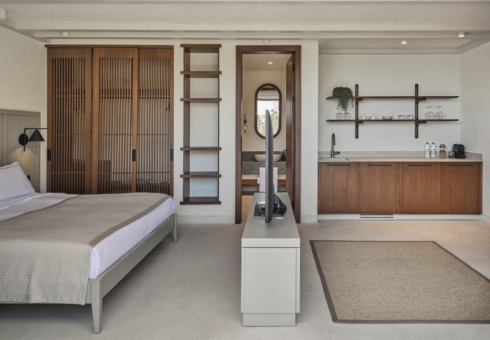 A modern bedroom with a bed, a bedside table, a wardrobe, a bathroom with a sink and mirror, and a shelf with decorative items.