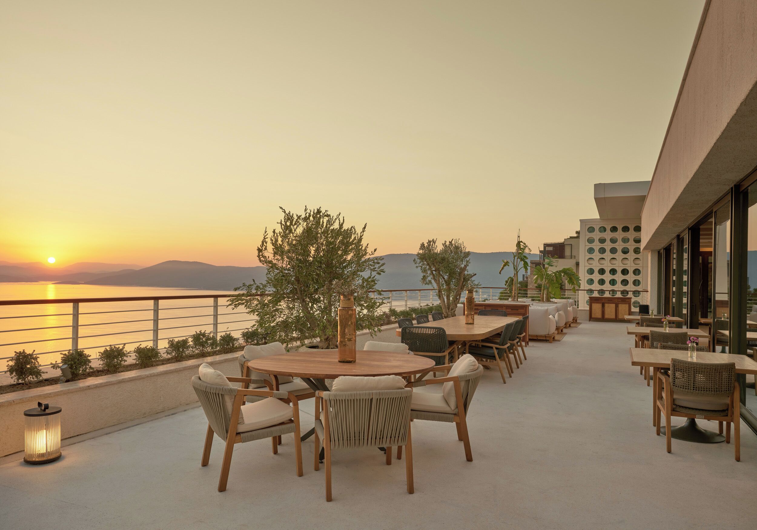 An outdoor seating area with round tables and chairs, overlooking a body of water and mountains.