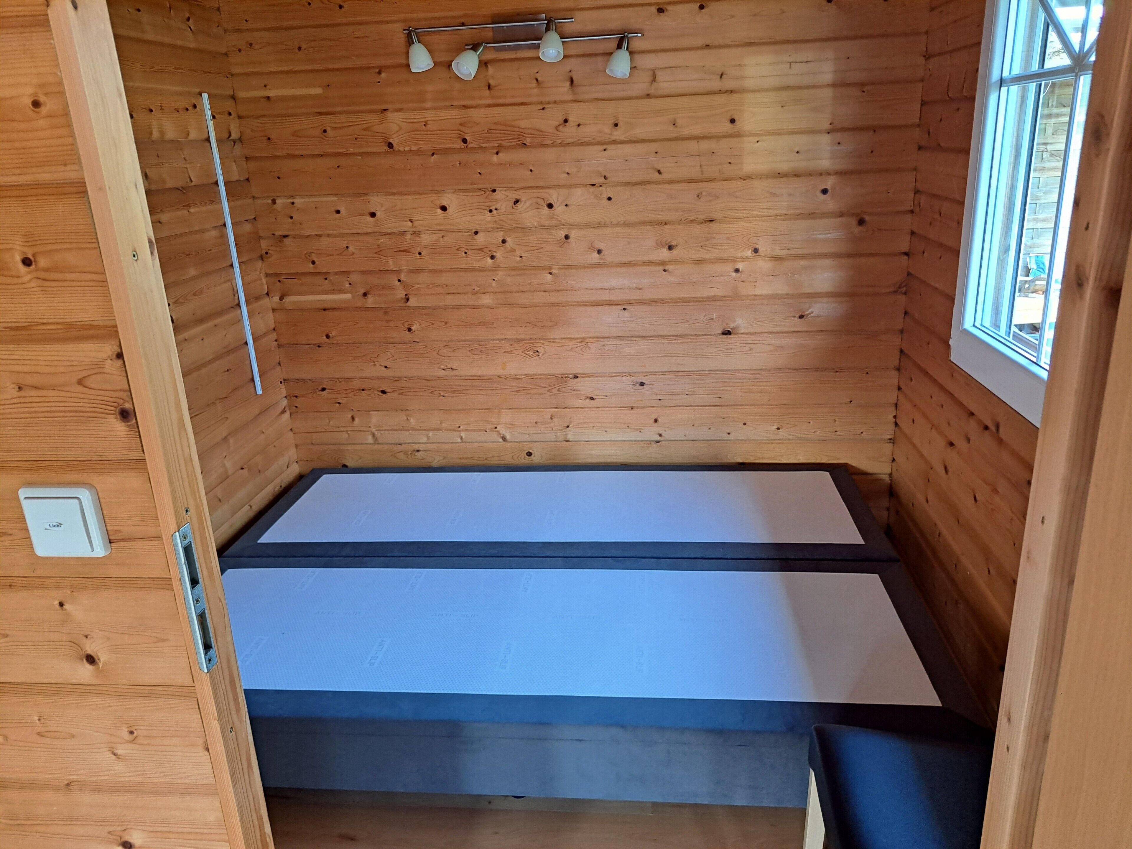 Une petite pièce en bois, aménagée comme une cabane, avec un lit, une fenêtre et un luminaire.