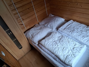 A wooden bunk bed with white bedding, a wooden wardrobe, and a metal ladder.