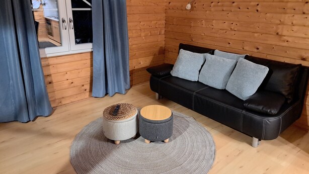 A wooden interior with a black leather sofa, grey cushions, and two woven stools.