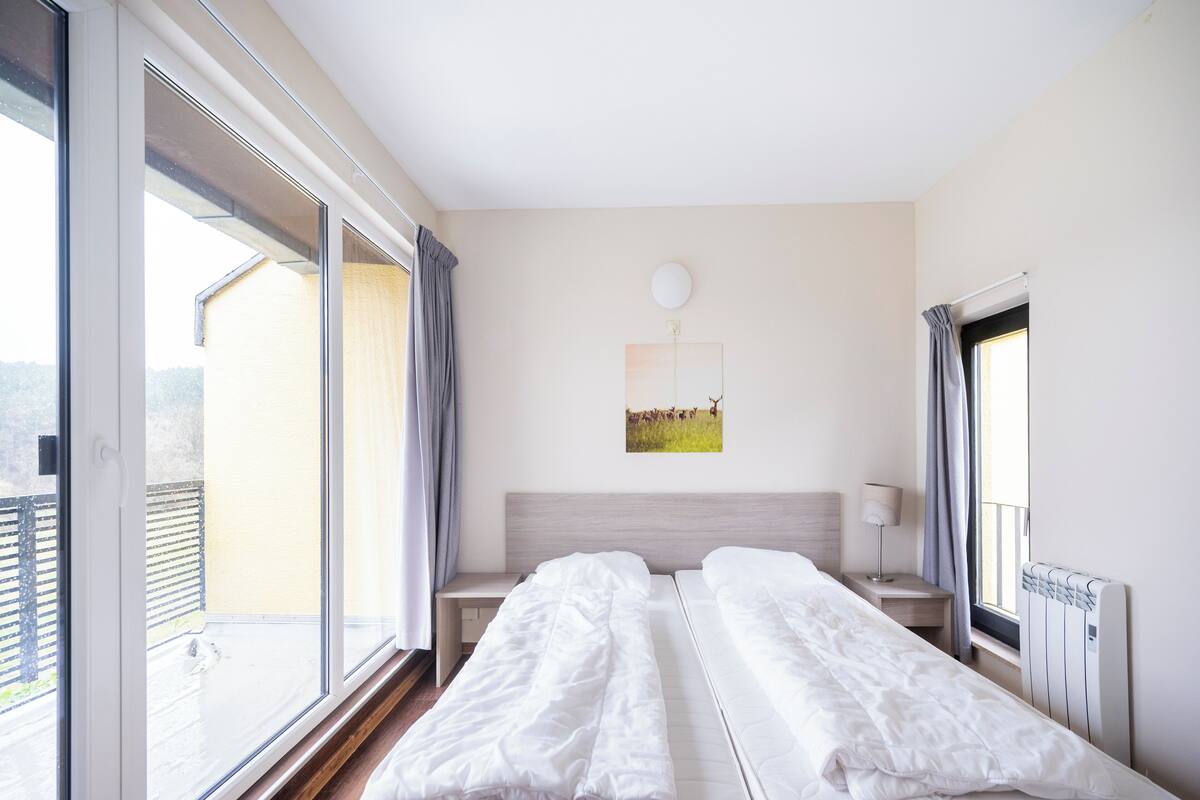 A hotel room with two beds, a window, a bedside table, a lamp, and a painting on the wall.
