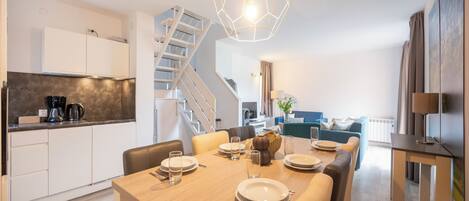 A modern kitchen with white cabinets, a dining area with a wooden table set for four, and a staircase leading to an upper level.
