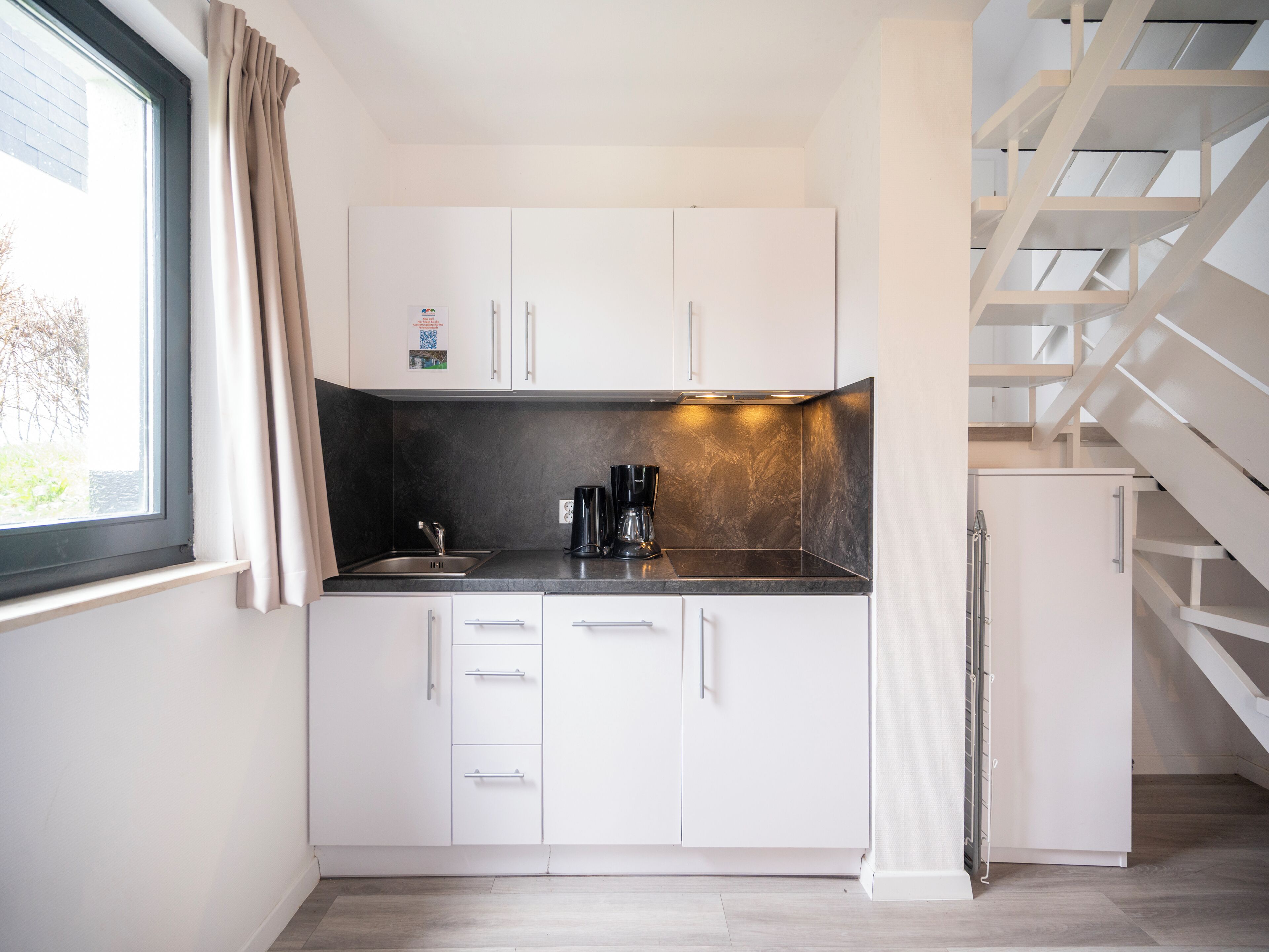 A compact kitchen with white cabinets, a black countertop, and a window with curtains.