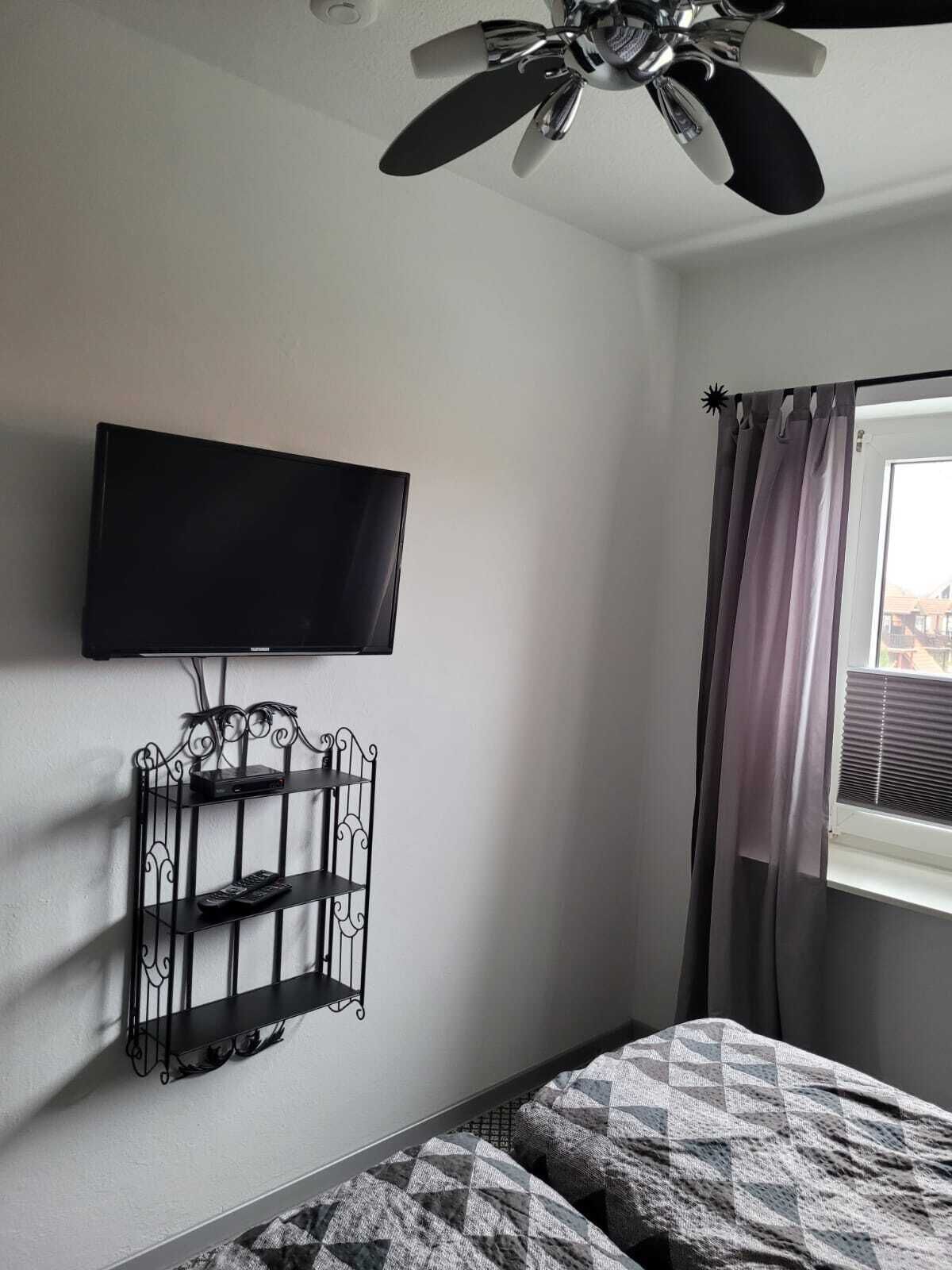 Un dormitorio con ventilador de techo, televisor montado, un mueble metálico para televisor, ventana con cortinas y una colcha estampada.