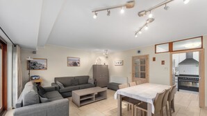 Un salon moderne avec un canapé gris, une table basse en bois et une table à manger avec des chaises. On aperçoit un coin cuisine avec une cuisinière et des placards, le tout visible à travers une porte ouverte.