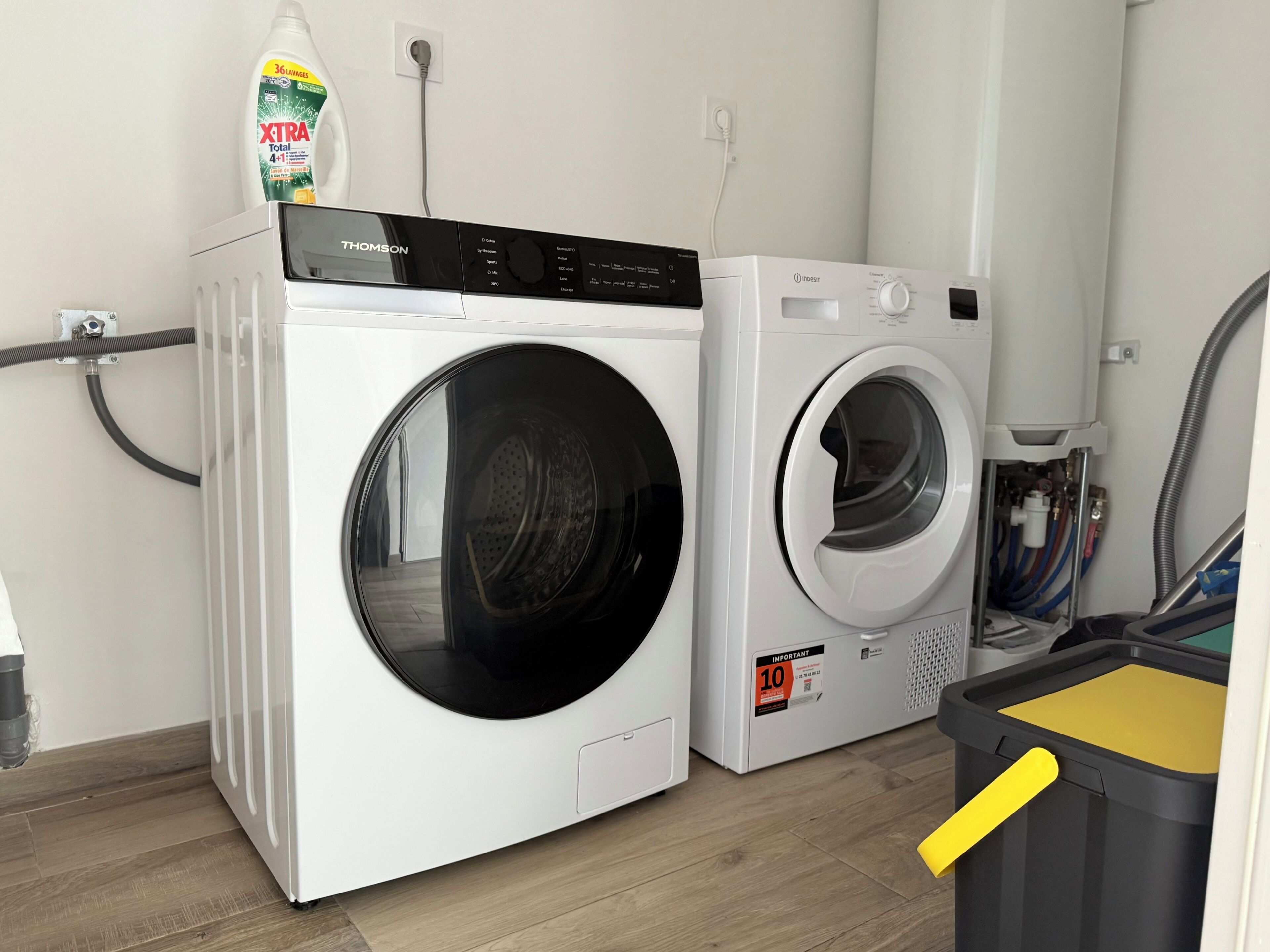 Un espace buanderie comprenant deux machines à laver à chargement frontal, un bidon de lessive et une poubelle.