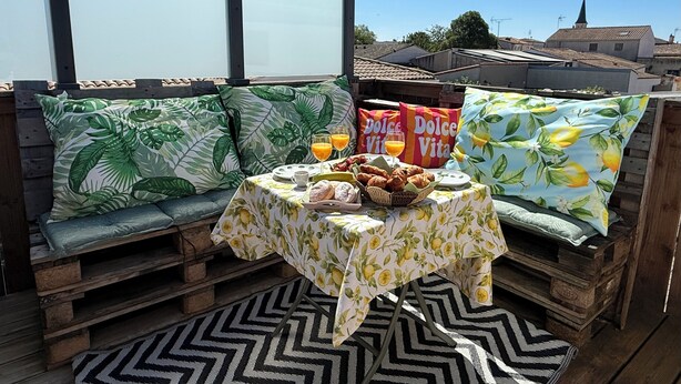 Een terras op het dak met een houten bankje, een tafel met een tafelkleed met fruitprint en twee kussens met een tropisch bladmotief.