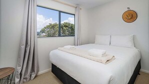 A bedroom with a large bed, a window with a view of trees, and a hat on the wall. - Beach House @ 406 - Matarangi (Matarangi)
