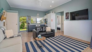 A spacious living room with a black leather sofa, a coffee table, and a striped rug. A kitchen area with a white cabinet and a flat-screen TV is visible in the background. - Beach House @ 406 - Matarangi (Matarangi)
