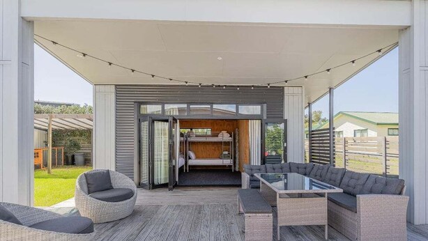 A modern outdoor living area with a glass table, wicker furniture, and a view of the backyard. - Beach House @ 406 - Matarangi (Matarangi)