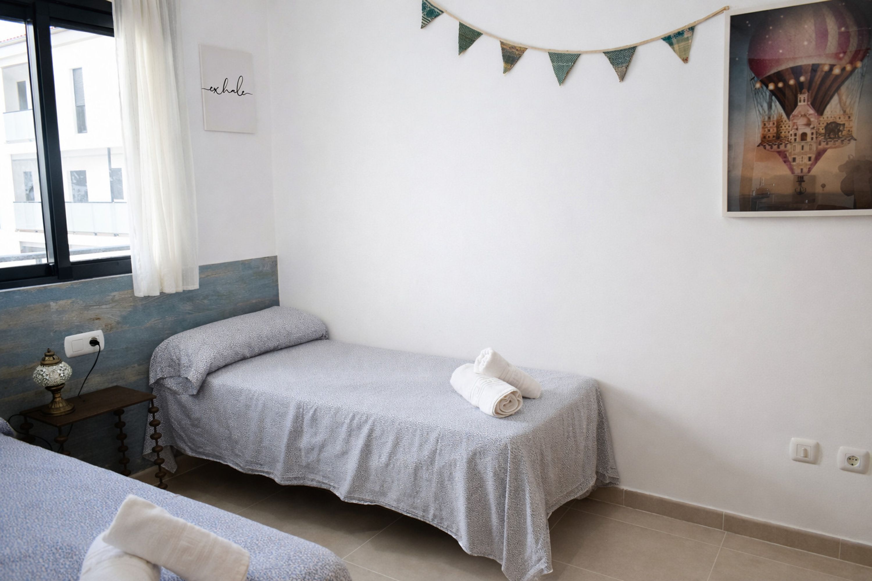 Una cama individual con una colcha gris y una toalla enrollada encima, una mesita de noche con una lámpara, y una ventana con cortinas.