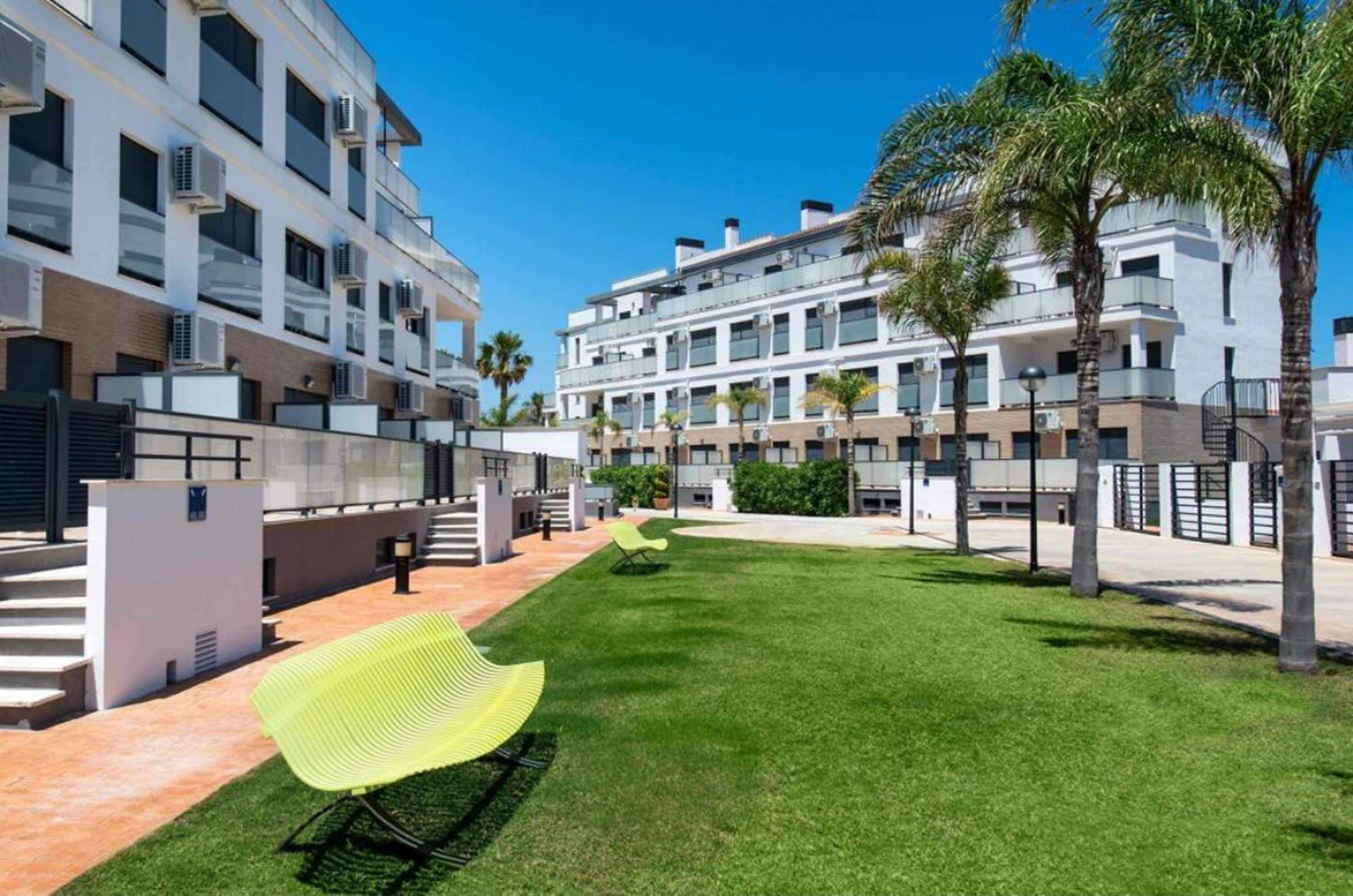 Un edificio moderno con varios balcones, un césped cuidado y algunas palmeras.