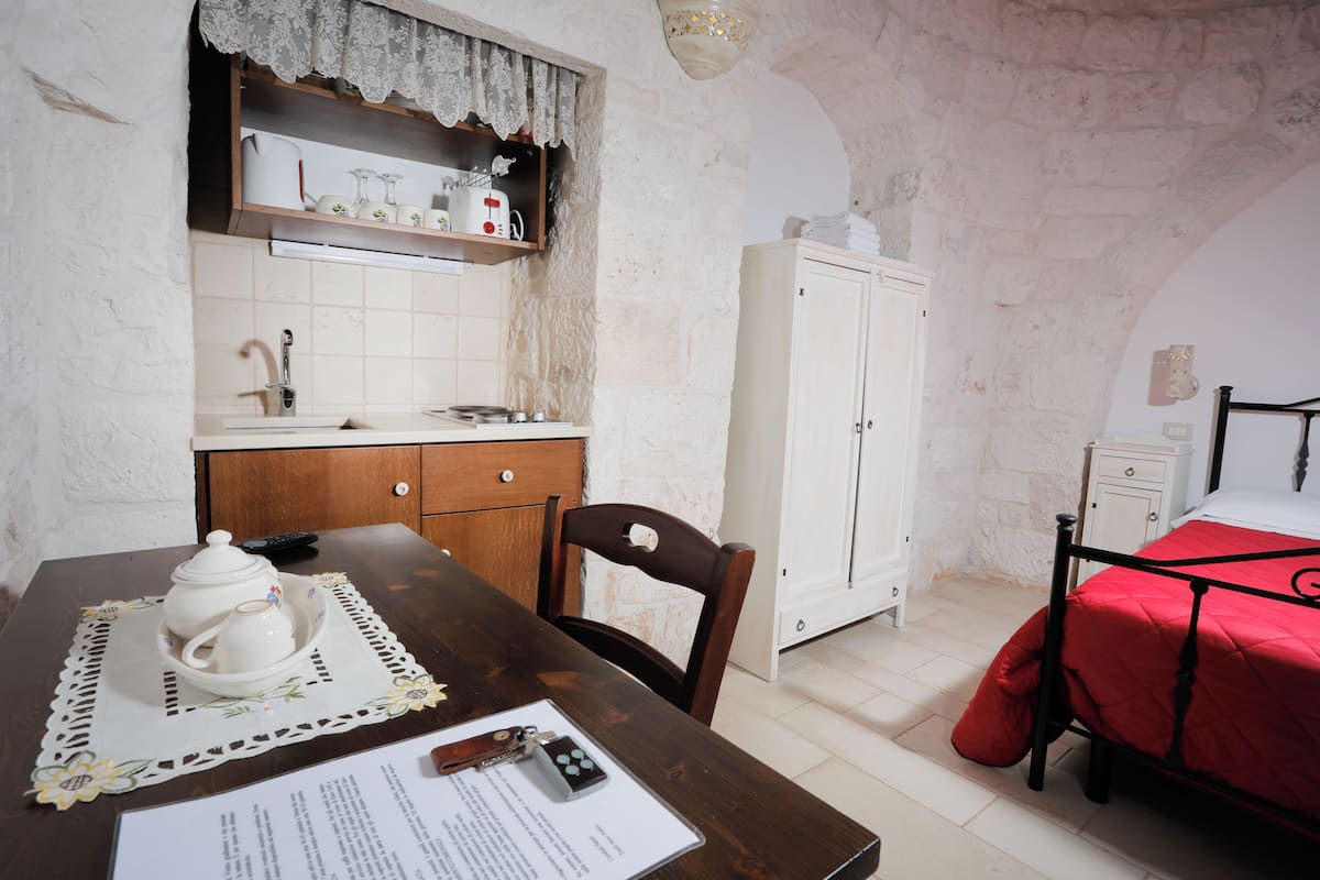 Une pièce avec une table en bois, une armoire blanche, un lit recouvert d’une couverture rouge et une garde-robe blanche.