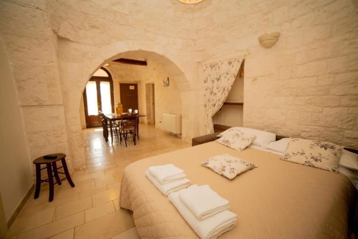 Une chambre avec un grand lit, un petit tabouret, une table en bois et une chaise.