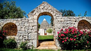 A stone archway leading to a building with a conical roof, surrounded by greenery and flowering plants. - Trulli Arco Antico (Locorotondo)