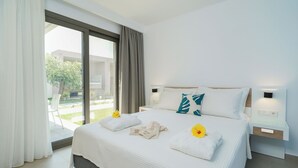 A modern hotel room with a large bed, white bedding, a view of the outdoors, and a nightstand with a lamp. - OLIVIA RESORT (Thasos)