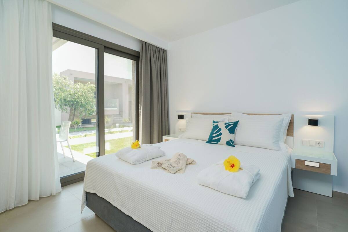 A modern hotel room with a large bed, white bedding, a view of the outdoors, and a nightstand with a lamp.