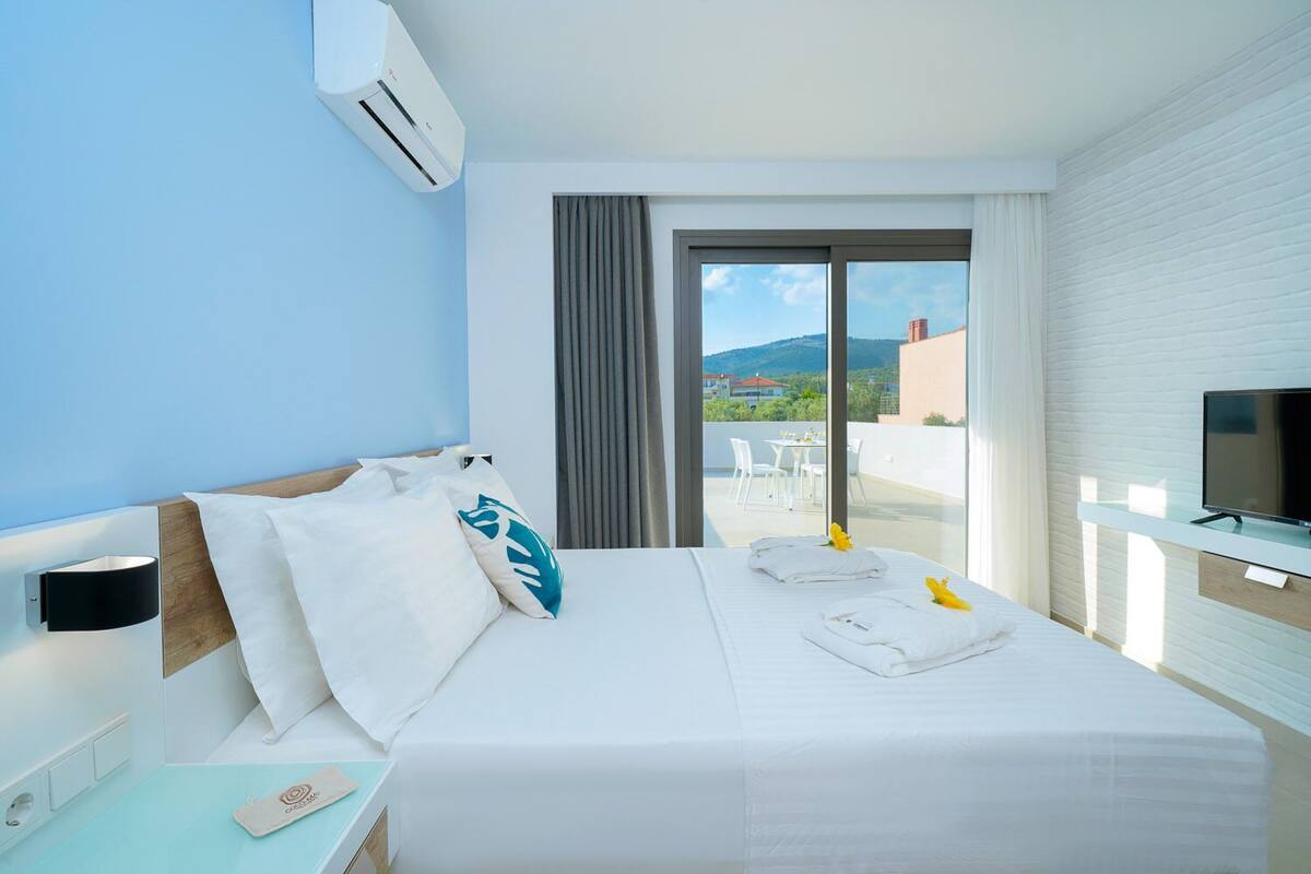 A hotel room with a bed, a TV, and a balcony with a view of buildings and hills.