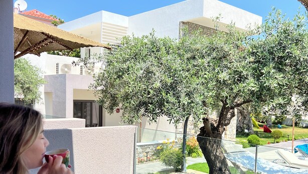 A balcony with a table, a person drinking, and a view of a garden with trees and a pool. - OLIVIA RESORT (Thasos)