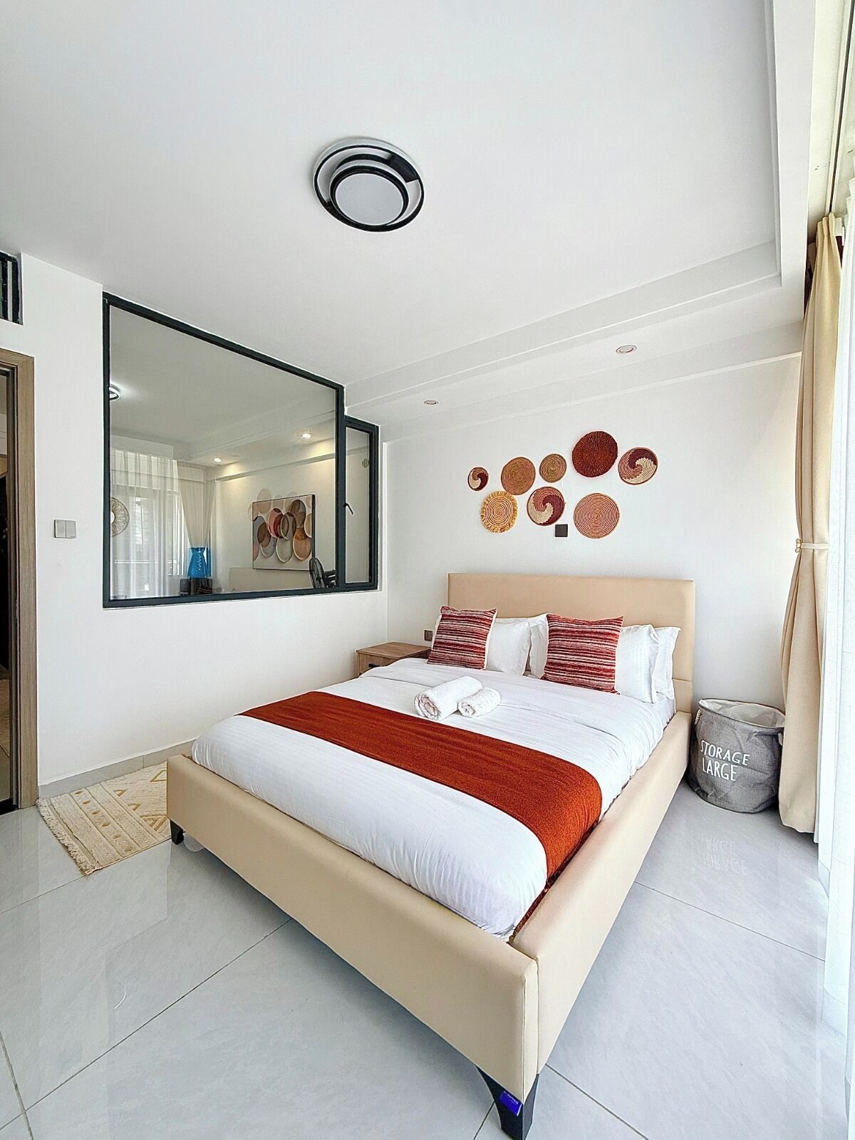 A modern hotel room with a large bed, a mirror, wall decor, and a storage bin.