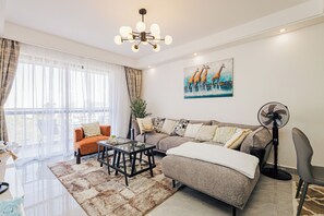 Un salon moderne avec un canapé d’angle, une table basse en verre et une table à manger. Il y a une grande fenêtre avec des rideaux légers, un tableau représentant des girafes accroché au mur et un ventilateur sur pied.
